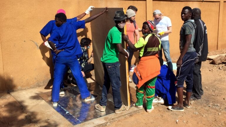 Eine Gruppe von Menschen arbeitet im Freien zusammen, um eine große rechteckige Platte anzuheben und zu positionieren, als Teil des Mini-Hybridkraftwerks für eine Krankenstation in Burkina Faso, einem Hilfsprojekt, an dem autarxia beteiligt war.