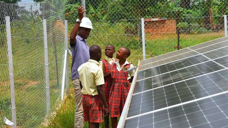 Ein Mann mit Schutzhelm erklärt drei Schulkindern in karierten Uniformen eine PV-Anlage, die an einem eingezäunten und begrünten Gelände steht - Teil eines Hilfsprojekts, an dem autarxia beteiligt war, zur Stromversorgung eines Waisenhauses in Uganda.
